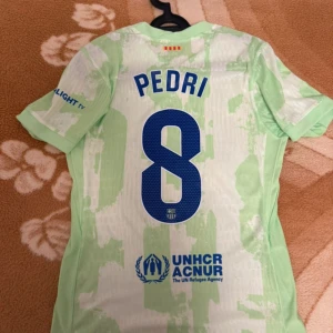 FC Barcelona Pedri fotbollströja Nike - Säljer en FC Barcelona fotbollströja med Pedri och nummer 8 på ryggen. Tröjan är ljusgrön med vita inslag, har klubbmärke och Spotify-logga på bröstet samt UNHCR-tryck bak. Kortärmad modell i lätt och ventilerande material, perfekt för match eller träning.