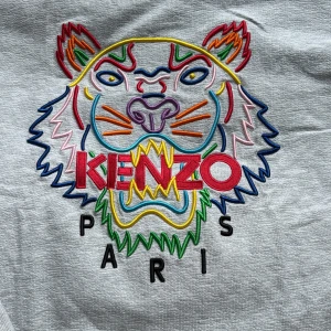 Grå Kenzo sweatshirt med tigerbrodyr - Säljer en grå sweatshirt från Kenzo med ikonisk färgglad tigerbrodyr och logga på bröstet. Tröjan har rund hals, långa ärmar och breda muddar. Perfekt statement-plagg för dig som gillar streetstyle och vill sticka ut.