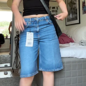 Blå denimshorts från Stradivarius - Säljer ett par blå denimshorts från Stradivarius i storlek 36. Modellen är loose fit med raka ben och klassisk femficksdesign. och är tillverkade i bomull med ljusblå tvätt och synliga sömmar. Perfekt för dig som gillar avslappnad stil.