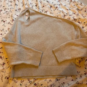 Grå stickad oneshouldertröja - Säljer en grå stickad tröja med oneshoulder-design och breda ribbade muddar vid ärmslut och nederkant. Tröjan har en avslappnad passform och är perfekt för kyligare dagar. Enkel och stilren look som passar till det mesta.