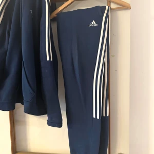 Blå Adidas hoodie med vita ränder - Säljer en marinblå Adidas hoodie med klassiska vita ränder längs ärmarna och Adidas-logga på bröstet. Tröjan har huva med snörning och en stor magficka. Perfekt för träning eller chill. Materialet är mjuk bomullsblandning.