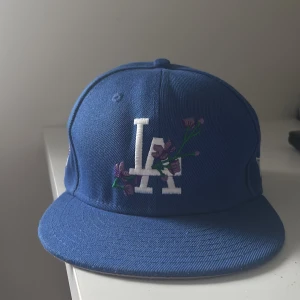 Blå LA keps från New Era 59FIFTY - Snygg blå keps från New Era 59FIFTY med LA-logga framtill och baktill. Framtill är loggan dekorerad med broderade lila blommor och gröna detaljer. Priset är inte hugget i sten vid snabbt köp! Hör av dig vid frågor