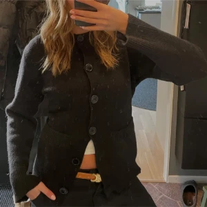 Svart stickad kofta med stora knappar - Mysig svart stickad kofta med stora knappar framtill och två fickor. Långärmad modell med ribbade muddar och klassisk rak passform. Perfekt att slänga över en t-shirt för en chill look.