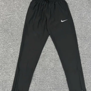 Svarta träningsbyxor från Nike - Svarta träningsbyxor från Nike med klassisk vit swoosh-logga på låret. Byxorna har elastisk midja med dragsko och är tillverkade i ett lätt och smidigt syntetmaterial. Perfekta för sport eller chill, med smal passform och enkel design. Det är storlek L men är liten i storleken och passar som M