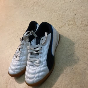 Vita och svarta Puma fotbollsskor - Säljer ett par klassiska vita och svarta Puma fotbollsskor med bruna sulor och blå detaljer. Skorna har snörning, rund tå och är tillverkade i syntetmaterial med quiltad ovandel. Perfekta för inomhusspel och snygga med den ikoniska Puma-loggan på hälen.
