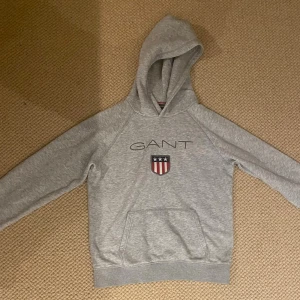 Grå hoodie från GANT med logga - Snygg grå hoodie från GANT med broderad logga och amerikansk sköld på bröstet. Tröjan har huva, magficka och ribbade muddar. Perfekt för chill dagar och enkel att matcha med jeans eller joggers.