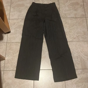 Gråa randiga kostymbyxor H&M 34 - Gråa kostymbyxor från H&M i storlek 34 med tunna vita ränder. Byxorna har hög midja och raka, lite vidare ben. 