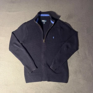Mörkblå stickad fullzip Polo Ralph Lauren - Snygg mörkblå stickad fullzip från Polo Ralph Lauren. Tröjan är i storlek Large. Tröjan är i ett jättebra skick och det finns inga tecken på användning. Skriv om du har några funderingar.