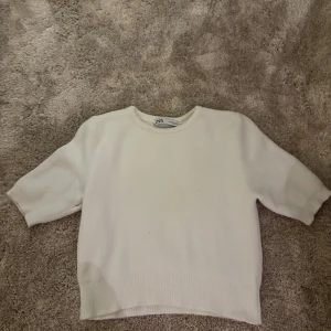Vit stickad tröja från Zara - Säljer en enkel och stilren vit stickad tröja från Zara i storlek M. Tröjan har rund halsringning, korta ärmar och ribbade detaljer vid hals, ärmslut och nederkant. Perfekt för dig som gillar minimalistisk och clean stil.