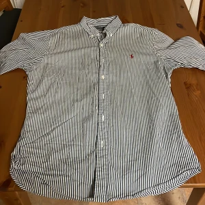 Randig skjorta Polo Ralph Lauren M - Slim fit skjorta från Polo Ralph Lauren i storlek M. Snyggt svartvit randigt mönster, klassisk krage och knappar framtill. Ikonisk röd logga broderad på bröstet. Tillverkad i mjuk bomull, perfekt för dig som gillar stilrena och tidlösa plagg.