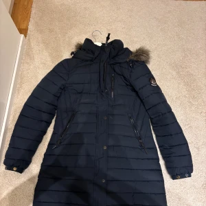 Mörkblå lång dunjacka Superdry - Säljer en mörkblå lång dunjacka från Superdry med huva och fuskpälsdetalj. Jackan har flera dragkedjefickor, knappar framtill och ett snyggt Superdry-märke på ärmen. Perfekt för kalla dagar med sin quiltade design och stilrena look.