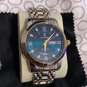 Poedagar herrklocka i silver och guld platerad  - Stilren herrklocka från Poedagar med blå urtavla, romerska siffror och datumvisning. Armbandet är i rostfritt stål med silver- och guldfärgade länkar. Klockan har guldfärgad krona och detaljer samt glittriga markörer på urtavlan rost fritt stål.