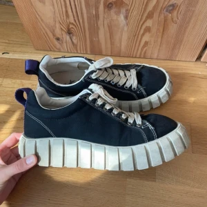 Chunky svarta sneakers med vit sula - Säljer ett par chunky svarta sneakers från Eytys med kraftig vit platt sula och vita snören. Skorna har en cool kontrast mellan svart textil och den grova sulan, samt en lila hälflik för extra detalj. Perfekta för dig som gillar statement-skor.