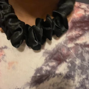Svart scrunchie-halsband i satin - Unikt svart halsband med scrunchie-look i glansigt satinmaterial. Halsbandet har en rynkad design som ger en cool och trendig vibe, och stängs med ett guldfärgat spänne. Perfekt accessoar för att lyfta din outfit med något annorlunda.