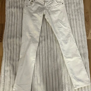 Vita jeansbyxor från Gina Jeans - Snygga vita jeansbyxor från Gina Jeans med raka ben och coola guldfärgade detaljer på bakfickorna. Klassisk femficksmodell med metallnitar och dragkedja. Perfekt för dig som gillar stilrena och trendiga jeans.