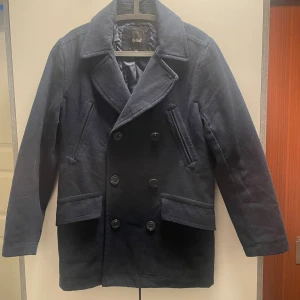 Kort Mörkblå skepparkavaj från deVal - Snygg mörkblå skepparkavaj från deVal i klassisk modell med dubbelknäppning och bred krage. Jackan har två sidofickor med lock och två snedställda bröstfickor. Tillverkad i ullblandning med slät fodrad insida för extra komfort.