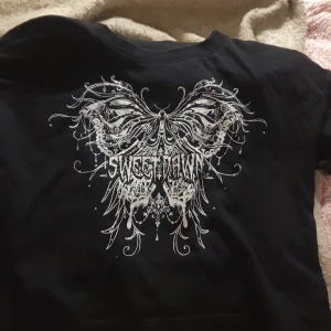 Svart croppad topp Sweet Dawn Lindex - Cool svart croppad topp från Lindex med stort vitt tryck framtill som föreställer fjärilsvingar och texten 'SWEET DAWN'. T-shirten har korta ärmar och rund halsringning. Perfekt för dig som gillar edgy och unik stil.