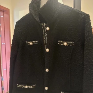 Svart teddyjacka med pärlknappar - Snygg svart teddyjacka med hög krage och fyra fickor framtill. Jackan har dekorativa pärlknappar och kontrastdetaljer i vitt vid fickor och knappslå. Perfekt för dig som gillar en chic och trendig look med lite extra detaljer.