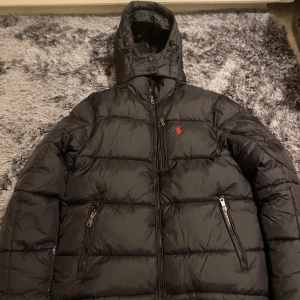 Svart pufferjacka från Polo Ralph Lauren - Svart pufferjacka från Polo Ralph Lauren med röd broderad logga på bröstet. Jackan har huva, två framfickor med dragkedja och quiltad design. Perfekt för kalla dagar och har en stilren look som funkar till det mesta.