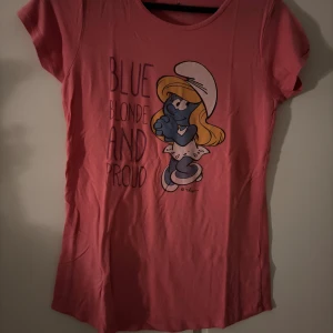 Rosa Smurfarna t-shirt med tryck - Cool rosa t-shirt med Smurfan-tryck och texten 'Blue Blonde And Proud' på framsidan. T-shirten har rund hals och korta ärmar, tillverkad i mjuk bomull. Perfekt för dig som gillar retro och tecknade figurer.