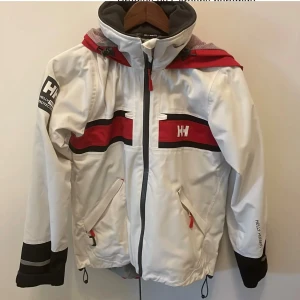 Helly Hansen vit och röd vindjacka - Snygg vindjacka från Helly Hansen i vitt med röda och svarta detaljer. Jackan har huva, dragkedja framtill, två fickor med dragkedja och logga på bröstet och ärmen. Perfekt för dig som gillar sportig stil och funktionella plagg.