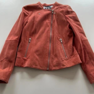 Korallfärgad skinnjacka med dragkedjor - Snygg korallfärgad skinnjacka med asymmetrisk dragkedja och två fickor med dragkedjor framtill. Jackan har en stilren design med lång ärm och markerade sömmar som ger en cool vibe. Perfekt för dig som vill sticka ut med färg och form.