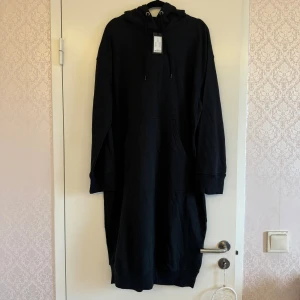 Svart hoodieklänning med huva - Svart hoodieklänning med lång ärm, huva och dragsko. Aldrig använd med prislapp på, köpt förra året. Klänningen har en stor känguruficka framtill och ribbade muddar vid ärmslut och nederkant. 🥰