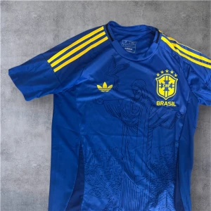 Blå Brasil Adidas fotbollströja - Säljer en blå Brasil fotbollströja från Adidas med gula detaljer och tryck. Tröjan har korta ärmar, gula ränder på axlarna och ett stort motiv av Kristusstatyn på framsidan. CBF-logga och Adidas-märke i gult. Perfekt för fans!