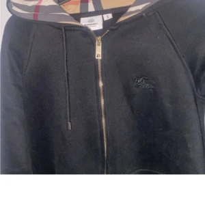 Svart Burberry hoodie med dragkedja - Svart hoodie från Burberry i storlek M med klassisk rutmönstrad huva, guldfärgad dragkedja och broderad logga på bröstet. Tröjan har snörning vid huvan och är tillverkad i mjuk bomull. Perfekt för dig som gillar stilrena designer med lyxig touch.