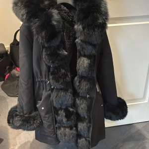 Svart parkajacka med pälsdetaljer - Parkajacka i svart med fluffiga pälsdetaljer runt huva, ärmslut och längs öppningen. Jackan är i storlek Xs-S och har en dragsko i midjan för att göra den mindre och mer formad, den har också stora fickor med knappar. Insidan är fodrad med mjuk päls för extra värme. Perfekt för kalla dagar, jackan går även att göra till en väst och är använd 2 vintrar och har endast några fläckar men går att skicka den till kemtvätt om man vill det. Pris går att diskuteras vid snabb affär. 