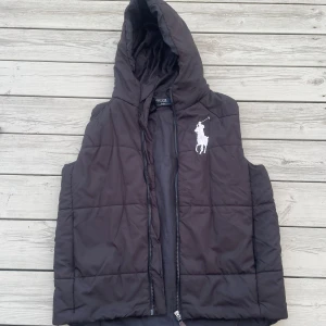 Svart dunväst med huva från Polo Ralph Lauren - Svart quiltad dunväst från Polo Ralph Lauren med stor vit broderad logga på bröstet. Västen har huva och dragkedja framtill, perfekt för lager-på-lager-stil. Klassisk och clean look som funkar till många outfits.