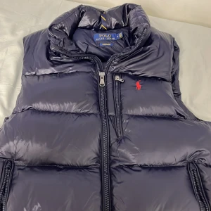 Mörkblå dunväst från Polo Ralph Lauren - Snygg mörkblå dunväst från Polo Ralph Lauren med glansig finish och klassisk röd logobrodering på bröstet. Västen har hög krage, två sidofickor med dragkedja och en robust tvåvägsdragkedja framtill. Perfekt för lager-på-lager under kyliga dagar.