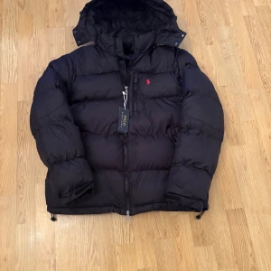Svart dunjacka från Polo Ralph Lauren - Svart dunjacka från Polo Ralph Lauren med huva, dragkedja och klassisk röd logga på bröstet. Jackan är tjockt vadderad och har justerbara muddar samt dragsko nedtill. Perfekt för kalla dagar och har en stilren look som funkar till allt.