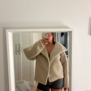 Beige kappa - Säljer en beige kappa med bred krage och asymmetrisk knäppning. Jackan har en loose passform och är tillverkad i mjukt fleece-liknande material. Perfekt för dig som gillar chill och comfy vibes. Långärmad och riktigt mysig att bära under kyliga dagar.