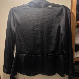 Svart omlottblus med knyt i siden - Snygg svart blus i siden med omlottdetalj och djup v-ringning framtill. Blusen har långa ärmar med knyt vid ärmslut och en liten ståkrage. Materialet är glansigt och ger en lyxig känsla. Perfekt för dig som gillar stilrena och trendiga plagg.