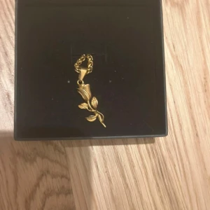 Guldigt halsband med ros - Snyggt halsband från Celett med en detaljerad ros som hänge. Kedjan och hänget är i guldig färg och har en cool, lite chunky kedja. Perfekt accessoar för att lyfta din outfit med en touch av blommig stil.