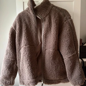 Beige teddyjacka från H&M XS - Mysig teddyjacka i beige från H&M, storlek XS. Jackan har hög krage, dragkedja framtill och två sidofickor. Materialet är fluffigt och ger en chill vibe, perfekt för kalla dagar. Passformen är normal och jackan har långa ärmar.
