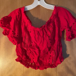 Röd croppad blus med spetsdetaljer - Säljer en röd croppad blus med korta puffärmar och snygga spetsdetaljer framtill. Blusen har en rynkad, elastisk kant vid halsringning och nederkant som ger en cool och feminin vibe. Perfekt för dig som gillar färg och detaljer!