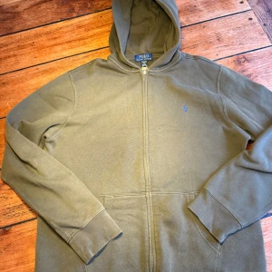 Olivgrön hoodie från Polo Ralph Lauren xs - Olivgrön hoodie från Polo Ralph Lauren i storlek xs. Klassisk design med dragkedja, huva och känguruficka. Broderad blå logga på bröstet. Perfekt för chill dagar och enkel att matcha med jeans eller joggers.