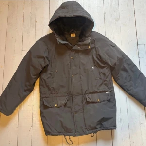 Carhartt svart vinterparkas med huva - varmoch fodrad - Snygg och varm parkas i svart, med huva och mjukt, varmt foder på insidan. Jackan har dragkedja framtill samt knappslå för extra vindskydd. Två stora fickor nedtill med lock och tryckknappar, samt två bröstfickor. Justerbar nederkant med dragsnöre. Jackan är använd men i gott skick och fungerar bra till vardag, vinterpromenader eller arbete utomhus. Levereras med dragkedja med en unik LEGO Darth Vader-nyckelring som dragkedjehänge (kan tas bort om man vill).
