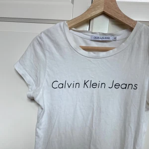 Vit t-shirt Calvin Klein Jeans XS - Vit t-shirt från Calvin Klein Jeans i storlek XS. Klassisk rund hals och korta ärmar, med svart logotyptryck framtill. Tillverkad i mjuk bomull. Använd ett fåtal gånger.