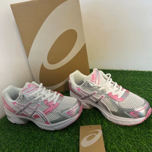 Asics GEL-1120 sneakers vit/rosa - Fräscha Asics GEL-1120 sneakers i vit mesh med detaljer i rosa och silver. Skorna har snörning, platt sula och rund tå. Snyggt sportig design med rosa inslag på sidor och häl, samt silverfärgade paneler. Perfekta för träning eller chill vardagsstil.