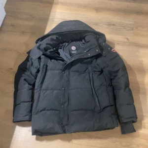 Svart pufferjacka från Canada Goose - Svart pufferjacka från Canada Goose med stor huva och klassisk logga på vänster ärm. Jackan har flera fickor med dragkedja och tryckknappar framtill. Perfekt för kalla dagar med sin tjocka, quiltade design och stilrena look.