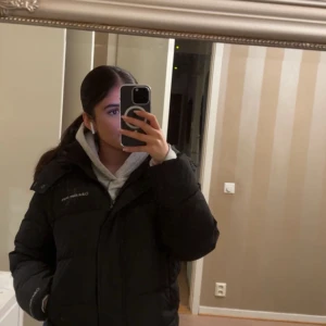 Svart pufferjacka från Calvin Klein - Svart pufferjacka från Calvin Klein med hög krage och huva. Jackan har en stilren design med logotyp på bröstet, dragkedja framtill och två sidofickor. Perfekt för kalla dagar och har en schysst, lite boxig passform som ger en cool vibe.