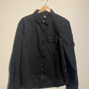 C.P. Company svart jacka, overshirt  - Svart overshirt från C.P. Company med klassisk krage och dragkedja framtill. Långärmad modell med ikonisk linsdetalj på vänster ärm. Tillverkad i bomull för en skön och stilren känsla.   Skick: Mycket gott skick, sparsamt använd.  Storlek: M men sitter som en S  Kolla gärna in mina andra annonser.   Skriv vid frågor eller ifall du vill ha fler bilder. 