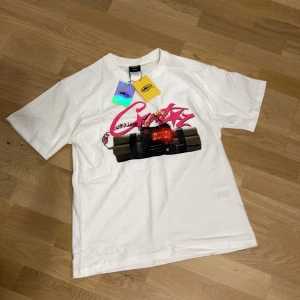 Vit t-shirt NO TIME 4 LUV från corteiz - Säljer en vit t-shirt från corteiz med tryck framtill av en digital bomb och graffiti-inspirerad text i rosa. På ryggen står det 'NO TIME 4 LUV' i stora svarta bokstäver. T-shirten har rund hals och korta ärmar. Materialet är mjuk bomull.