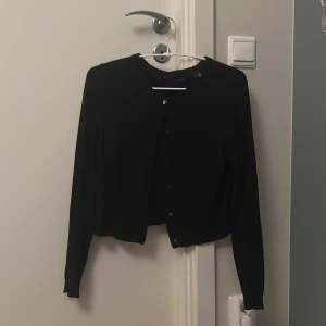 Svart kofta - En klassisk svart kofta från Vero Moda i storlek S. Koftan har rund halsringning, långa ärmar och knäppning framtill med små svarta knappar. Perfekt att slänga över en t-shirt eller linne för en enkel och stilren look.