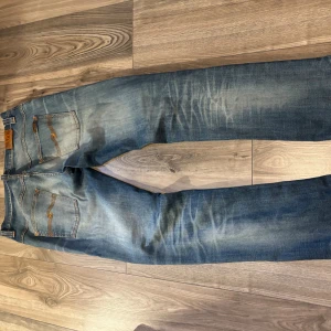 Nudie Jeans blå jeansbyxor W33 L34 - Snygga blå jeans från Nudie Jeans med klassisk femficksdesign och orangea sömmar. Har lite slitningar på framsidan men det ger också en karaktär. Jeansen har en rak passform och är tillverkade i slitstarkt bomullstyg. Fint detaljerade bakfickor med Nudie-logga och läderpatch i midjan. Perfekta för en avslappnad stil. Hör av dig vid frågor!!!