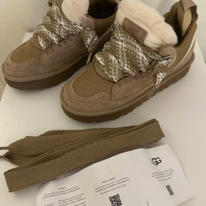 UGG lowmel - Snygga sneakers från UGG i beige mocka och textil med fluffig fårull på insidan. Skorna har breda, mönstrade snören i beige och vitt samt platt sula och rund tå. Perfekta för dig som vill ha både stil och komfort under höst och vinter.
