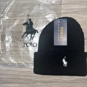 Svart ribbad mössa från Polo Ralph Lauren - Oanvänd svart ribbstickad mössa från Polo Ralph Lauren med klassisk uppvikt kant och vit broderad logga framtill. Snygg och enkel design som funkar till alla jackor. Materialet är mjukt och värmande, perfekt för kyliga dagar. 